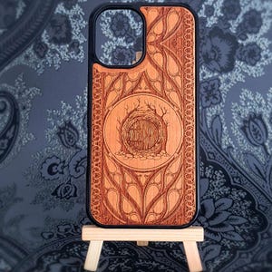 May include: A wooden phone case with a detailed, laser-etched design. The case features a central circular image of a hobbit hole, surrounded by intricate patterns and a black border. The case is displayed on a small wooden easel.
