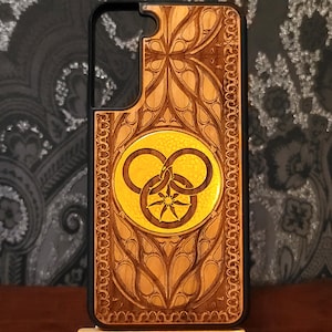 May include: A wooden phone case with a black border. The case features an intricate, carved design with a central yellow circle containing a symbol of three interlocking rings and a star. The wood has a light brown color.