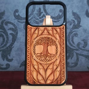 May include: A black phone case featuring a light brown wooden inlay. The wood surface is laser-engraved with a detailed circular design of a tree with intertwined roots and branches, surrounded by intricate decorative patterns on the sides.