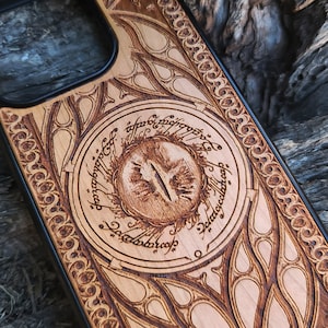 Könnte beinhalten: Eine Holz-Handyhülle mit einer detaillierten Schnitzerei des Auges von Sauron aus Der Herr der Ringe. Die Inschrift um das Auge lautet: "Ein Ring, sie alle zu knechten, Ein Ring, sie zu finden, Ein Ring, sie alle zu binden, und in der Finsternis sie zu fesseln."