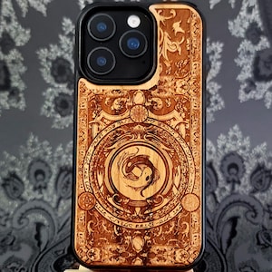 May include: A wooden phone case with a detailed engraved design. The case features a central circular emblem with two fish, surrounded by intricate patterns and text. The case is brown with a black border.