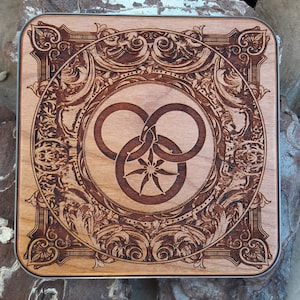 Puede incluir: Un posavasos de madera con un dise&ntilde;o intrincado tallado que presenta un s&iacute;mbolo estilizado del &aacute;rbol de la vida en el centro. El posavasos tiene un acabado marr&oacute;n oscuro y una forma cuadrada.