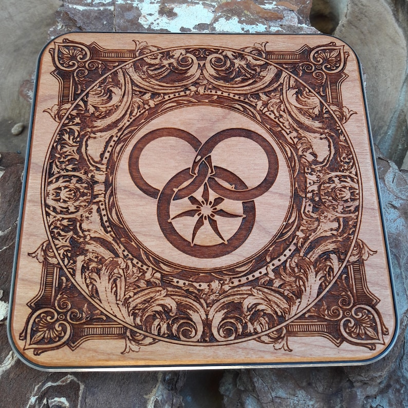 Puede incluir: Una placa de madera con un dise&ntilde;o intrincado y ornamentado. El centro presenta un s&iacute;mbolo estilizado de tres c&iacute;rculos entrelazados con una estrella en el centro.