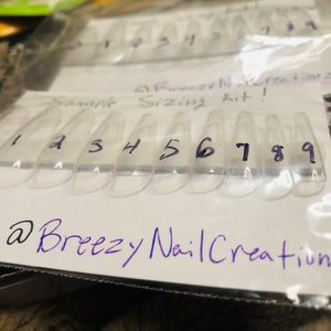 以下が含まれることがあります： 透明なプラスチック製のネイルサイズキットで、各ネイルに0から9までの番号が付けられています。キットは透明なプラスチック製の袋に梱包されており、紫色のインクで「@BreezyNailCreationz」という文字が書かれています。