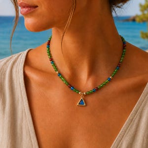 Puede incluir: Un collar de cuentas con pequeñas esferas verdes, azules y rojo oscuro intercaladas con detalles dorados. El collar incluye un colgante triangular dorado en el centro con una incrustación lisa de color azul intenso.