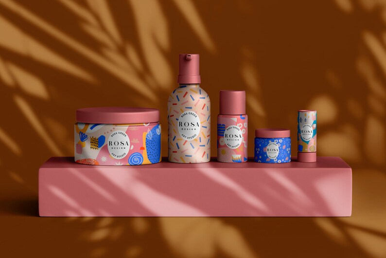 May include: A set of five colourful skincare products with a geometric pattern and the text "ROSA DESIGN ART STUDIO" on each. The products include a jar of cream, a bottle of lotion, a smaller bottle of serum, a jar of face mask, and a lip balm.