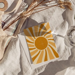 May include: A card with a retro sunburst design in shades of yellow and orange. The sunburst is centered on a large, yellow sun with a glittery texture. The card is lying on a white linen fabric with a glass of water and a woven bag in the background.