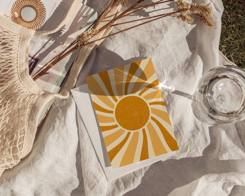 May include: A card with a retro sunburst design in shades of yellow and orange. The sunburst is centered on a large, yellow sun with a glittery texture. The card is lying on a white linen fabric with a glass of water and a woven bag in the background.