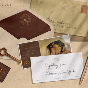 May include: A vintage postcard with a handwritten message that reads "Greeting from Queens, New York!" The postcard is sitting on a brown surface with a key, a brown envelope, and a pen.