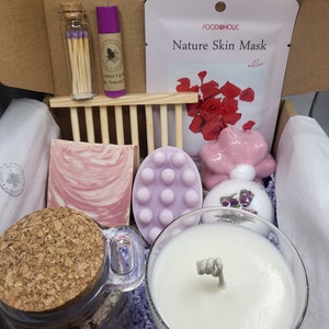 May include: A self-care gift box filled with various products. Includes a candle, bath bombs, soap, a face mask, matches, and lip balm. The box is brown with the word "WORKSHOP" printed on it.