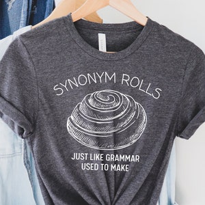 May include: A dark gray t-shirt with a white graphic of a cinnamon roll and the text "Synonym Rolls Just Like Grammar Used To Make".