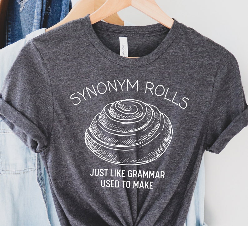 May include: A dark gray t-shirt with a white graphic of a cinnamon roll and the text "Synonym Rolls Just Like Grammar Used To Make".