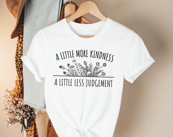 May include: White t-shirt with black text that reads "A little more kindness, a little less judgement". The text is centred on the shirt and is surrounded by a line of small flowers.