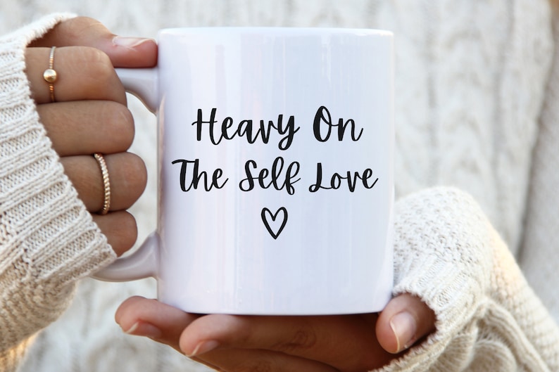 K&ouml;nnte beinhalten: Wei&szlig;e Keramiktasse mit schwarzem Text, der "Heavy On The Self Love" lautet, mit einem kleinen Herzsymbol unter dem Text.