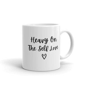 K&ouml;nnte beinhalten: Wei&szlig;e Keramiktasse mit schwarzem Text, der "Heavy On The Self Love" lautet, mit einem kleinen Herzsymbol unter dem Text.