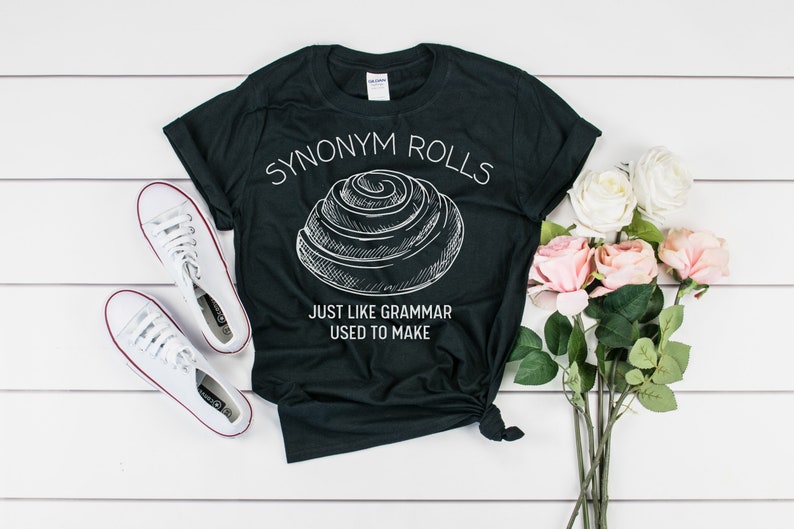 May include: Black t-shirt with white text that reads "Synonym Rolls Just Like Grammar Used To Make". A white outline drawing of a cinnamon roll is above the text.