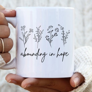 May include: White ceramic mug with a black line drawing of four different flowers and the text "abounding in hope".