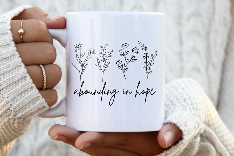 May include: White ceramic mug with a black line drawing of four different flowers and the text "abounding in hope".