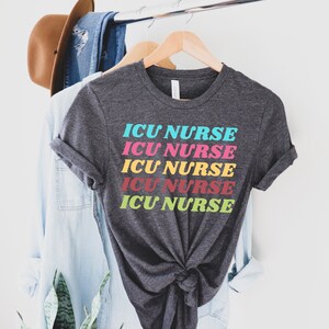 K&ouml;nnte beinhalten: Ein dunkelgraues T-Shirt mit dem Text "ICU NURSE", der f&uuml;nfmal in Regenbogenfarben wiederholt wird. Der Text ist vertikal gestapelt und jedes Wort hat eine andere Farbe.