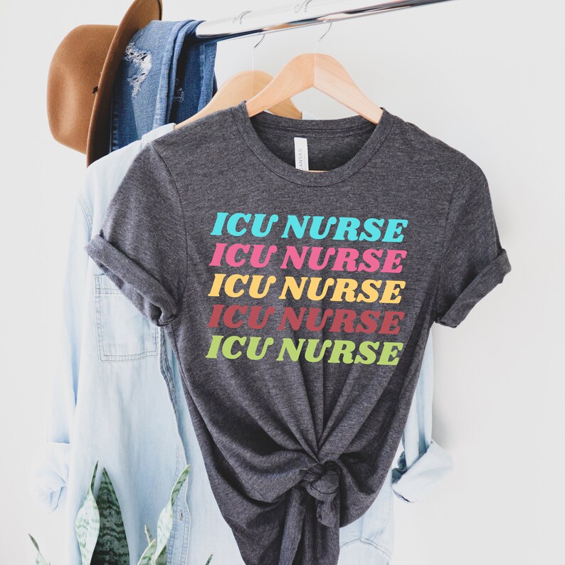 K&ouml;nnte beinhalten: Ein dunkelgraues T-Shirt mit dem Text "ICU NURSE", der f&uuml;nfmal in Regenbogenfarben wiederholt wird. Der Text ist vertikal gestapelt und jedes Wort hat eine andere Farbe.