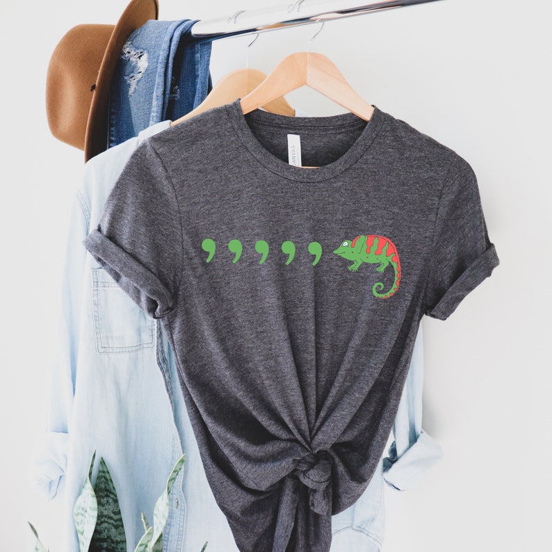 May include: A dark gray t-shirt with a green chameleon and five green quotation marks printed on the front.