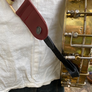 May include: A close-up of a banjo strap with a burgundy leather end and a black nylon strap. The strap is attached to a gold-colored banjo.