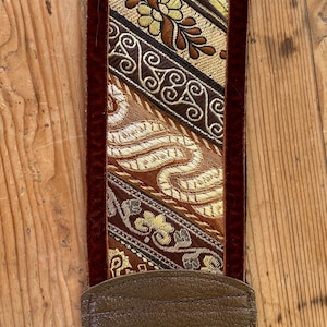 Może przedstawiać: Pasek do gitary z dekoracyjnym, tkanym wzorem w kolorach złotym, brązowym i kremowym. Pasek ma ciemnobrązowy skórzany koniec i ciemnoczerwony aksamitny brzeg.