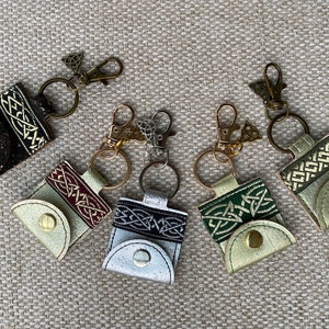 Könnte beinhalten: Sechs Schlüsselanhänger mit kleinen Ledertaschen und keltischen Knotendesigns. Die Taschen sind in Schwarz, Rot, Weiß und Grün mit goldenen Akzenten. Die Schlüsselanhänger sind an goldfarbenen Metallringen mit einem keltischen Knotenanhänger befestigt.