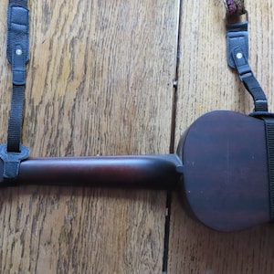 May include: A dark brown ukulele with black straps attached to the body and neck. The ukulele is lying on a wooden surface.