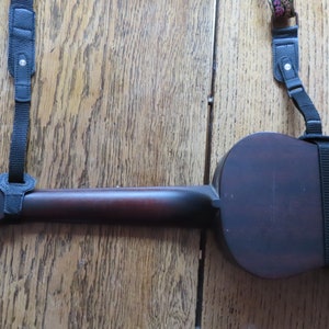 May include: A dark brown ukulele with black straps attached to the body and neck. The ukulele is lying on a wooden surface.