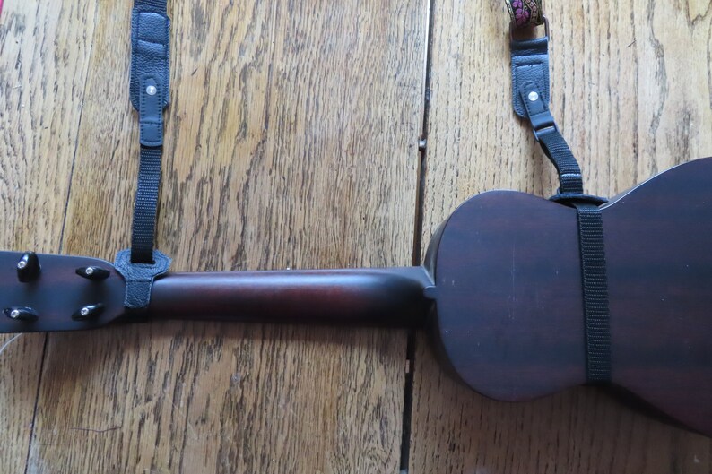May include: A dark brown ukulele with black straps attached to the body and neck. The ukulele is lying on a wooden surface.