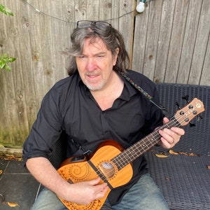 May include: A man wearing a black shirt and jeans is playing a brown ukulele with a decorative design. The ukulele has a black strap and a black pick-up.
