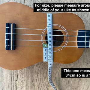 May include: A brown ukulele with white strings and a measuring tape wrapped around the body. The tape reads 34cm, indicating the size of the ukulele. The text "This one measures 34cm so is a S/M" is written below the ukulele.