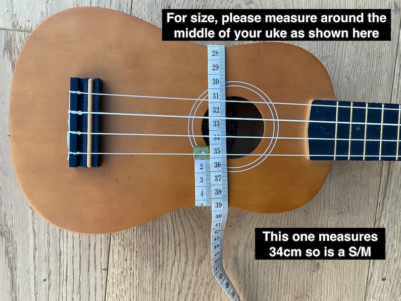 May include: A brown ukulele with white strings and a measuring tape wrapped around the body. The tape reads 34cm, indicating the size of the ukulele. The text "This one measures 34cm so is a S/M" is written below the ukulele.
