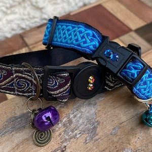 May include: A decorative pet collar with a blue Celtic knot design and a black buckle. The collar features a purple bell charm and a teal bell charm. The collar is made of fabric and has a black plastic buckle.