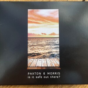 Puede incluir: Una caja de CD con fondo negro y una imagen rectangular de una puesta de sol sobre el océano. La imagen incluye un muelle de madera. El texto "PAXTON & MORRIS" y "is it safe out there?" están impresos en blanco.