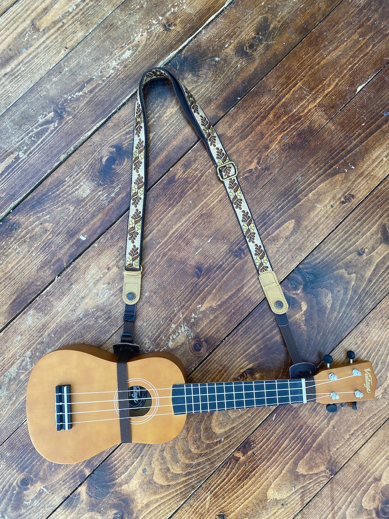 May include: A brown ukulele with a brown and white patterned strap. The ukulele has a black fretboard and a white string.