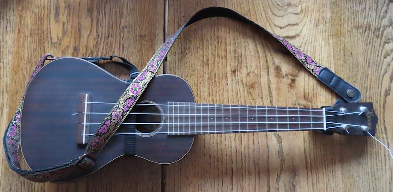May include: A brown ukulele with a floral patterned strap. The ukulele has a black strap attached to the body and a white string.