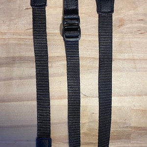 May include: Two black adjustable straps with leather accents and metal buckles. The straps are made of a woven material and have a textured finish.