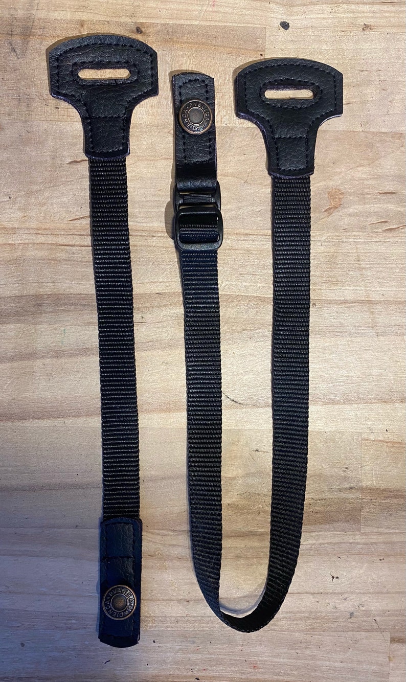 May include: Two black adjustable straps with leather accents and metal buckles. The straps are made of a woven material and have a textured finish.