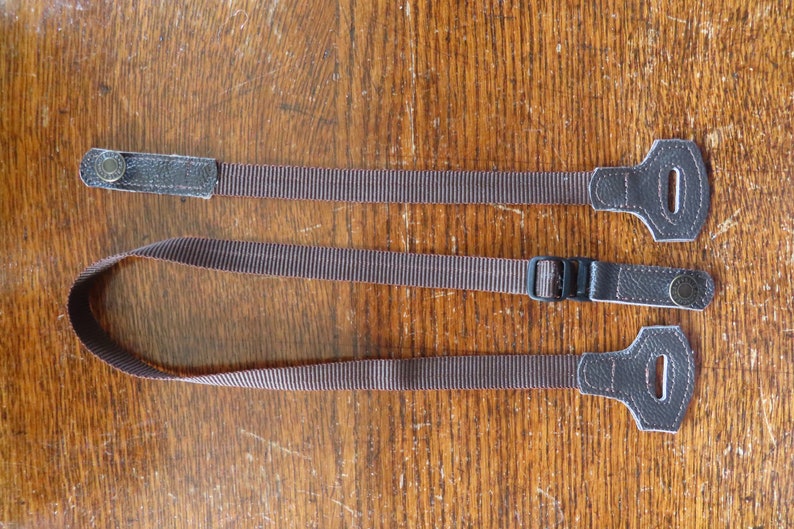 May include: Two brown leather camera straps with adjustable buckles. One strap has a metal button and the other has a metal ring.