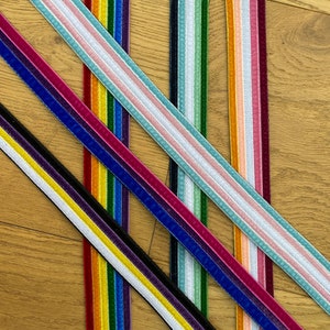 Pode incluir: Conjunto de fitas de veludo coloridas com acabamento branco. As fitas estão dispostas em forma de cruz sobre uma superfície de madeira.