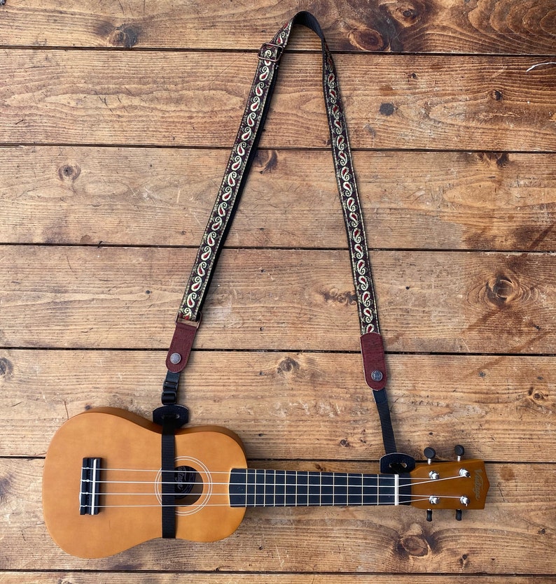 May include: A brown ukulele with a paisley patterned strap. The ukulele is on a wooden surface.