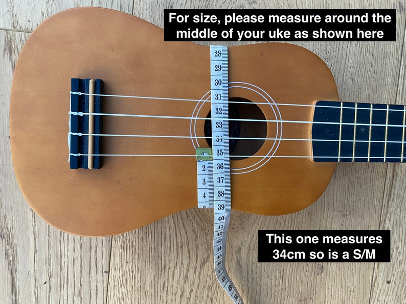 May include: A brown ukulele with white strings and a measuring tape wrapped around the body. The tape reads 34cm, indicating the size of the ukulele. The text "This one measures 34cm so is a S/M" is written below the ukulele.