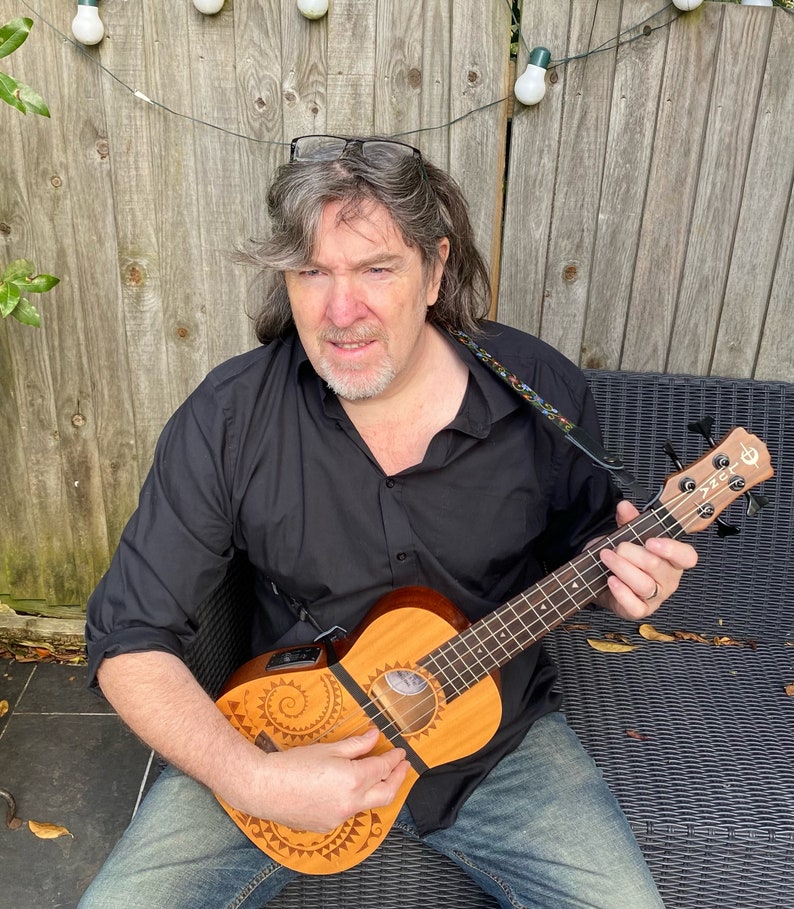 May include: A man sits in a chair holding a ukulele with a brown wooden body and a black fretboard. The ukulele has a decorative design on the body. The man is wearing a black shirt and blue jeans.
