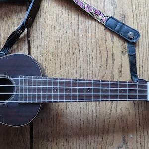 May include: A dark brown ukulele with a black strap and white strings. The ukulele has a floral patterned strap attached to it.