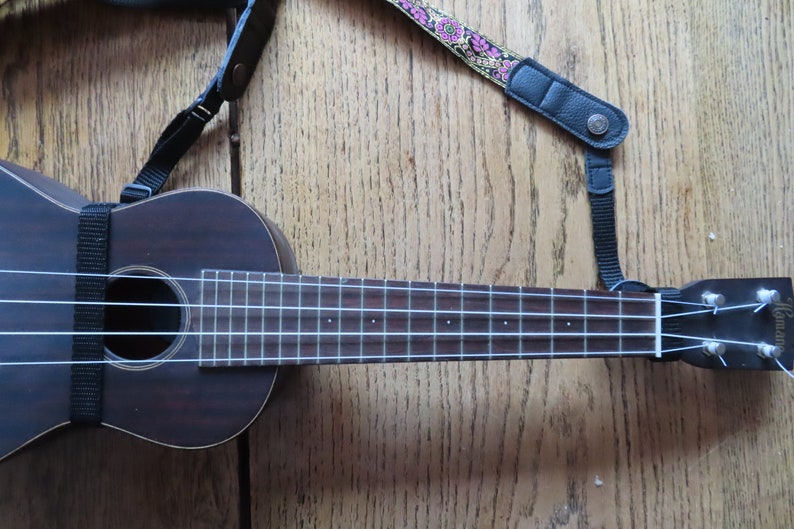 May include: A dark brown ukulele with a black strap and white strings. The ukulele has a floral patterned strap attached to it.