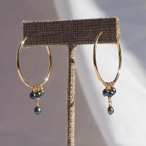 May include: A pair of gold hoop earrings, each featuring a cluster of three small, dark gray, teardrop-shaped pearls suspended from the bottom of the hoop. The earrings are displayed on a textured, brown, T-shaped jewelry stand.