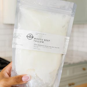 Op de afbeelding: Een doorzichtige plastic zak met Wagyu-rundertalg. Het etiket luidt "Wagyu Beef Tallow" en "Pure Rendered Grass Fed Beef Tallow". De zak wordt in een hand gehouden, met een lichte achtergrond.