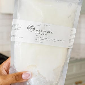 Puede incluir: Una bolsa de plástico transparente que contiene sebo de res Wagyu. La etiqueta dice "Wagyu Beef Tallow" y "Pure Rendered Grass Fed Beef Tallow". La bolsa se sostiene en una mano, con un fondo claro.